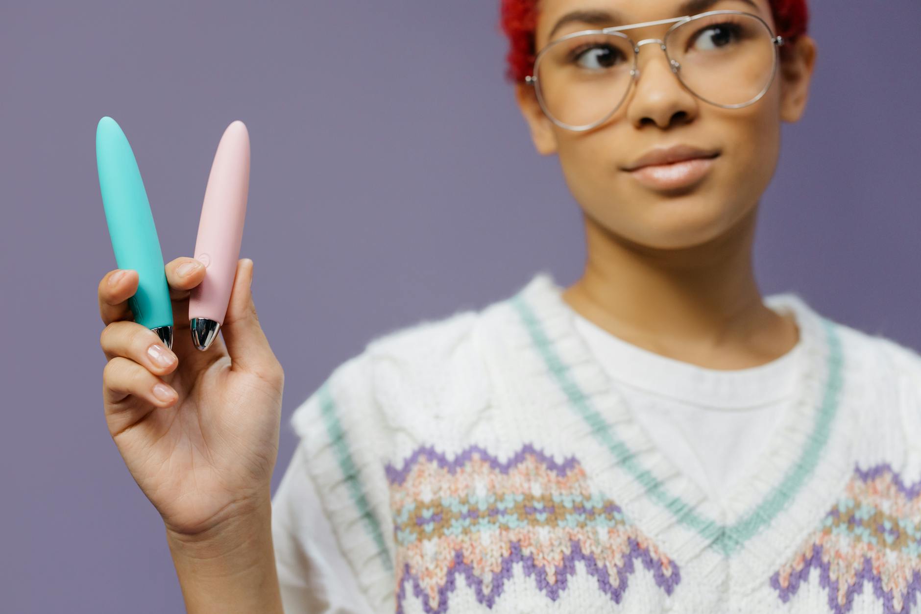 A young woman holds two colorful vibrators against a purple background.