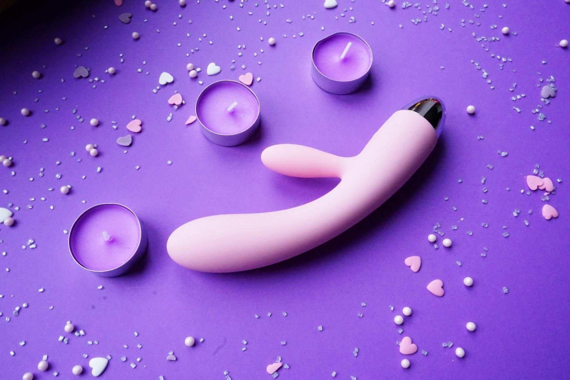 A pink clitoral vibrator with candles and heart confetti on purple background, symbolizing reconnection and intimate pleasure